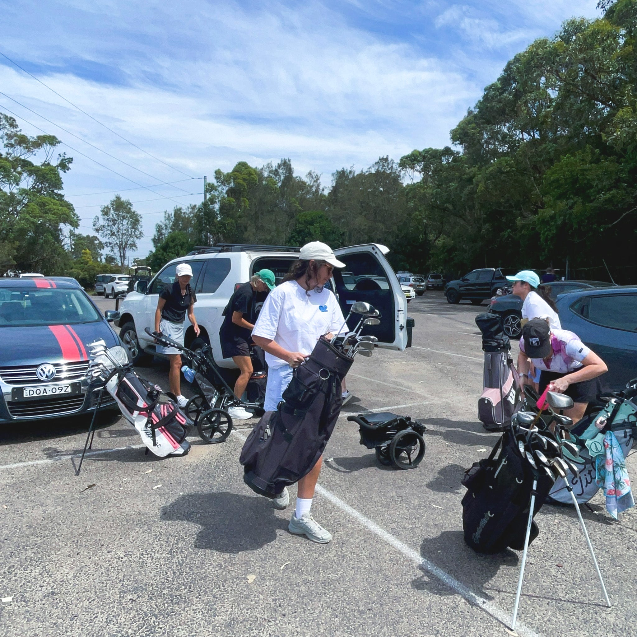 People with golf bags and equipment in a parking lot with cars and trees in the background