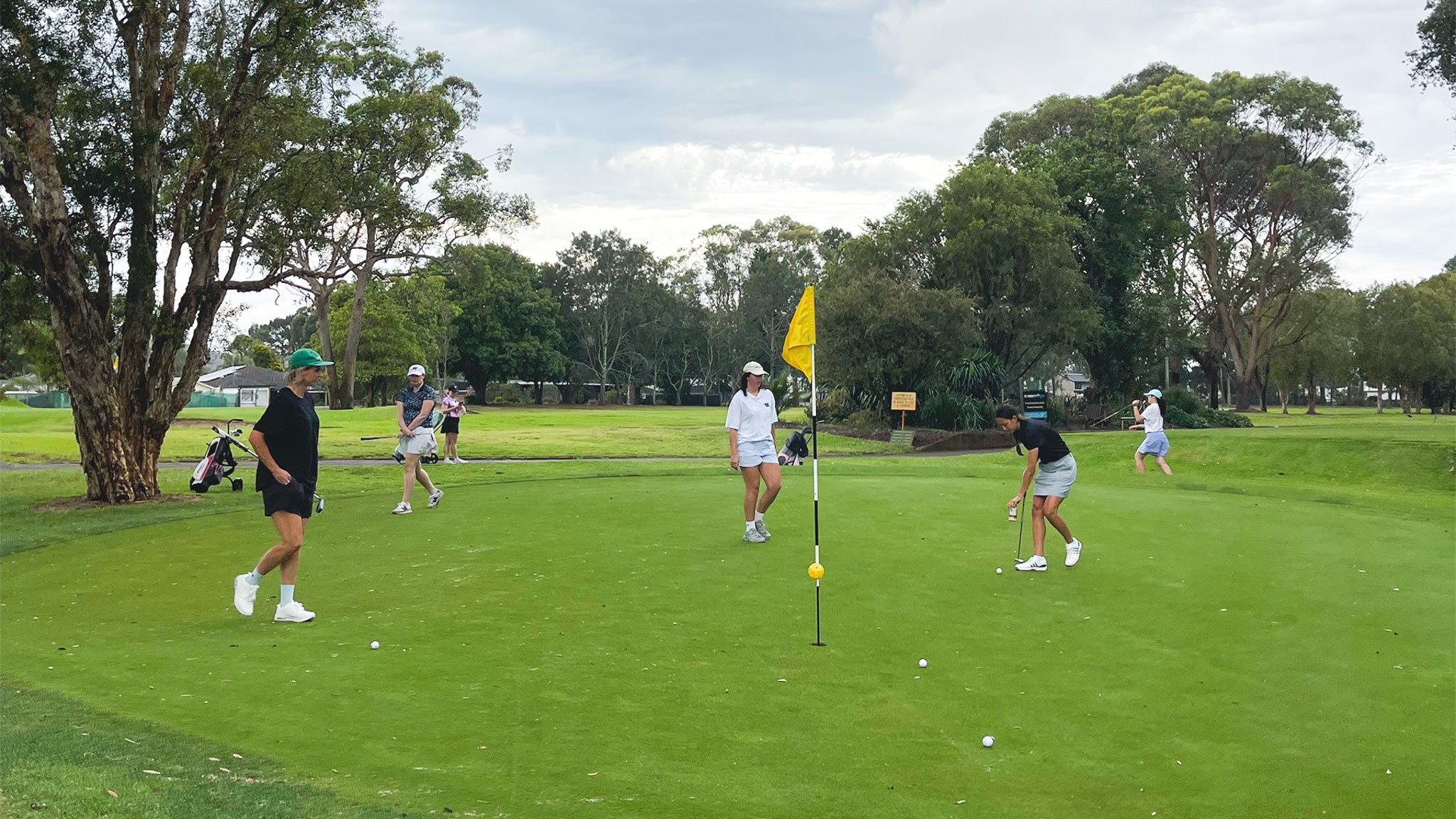 Golfers on a putting green with trees and a clear sky.