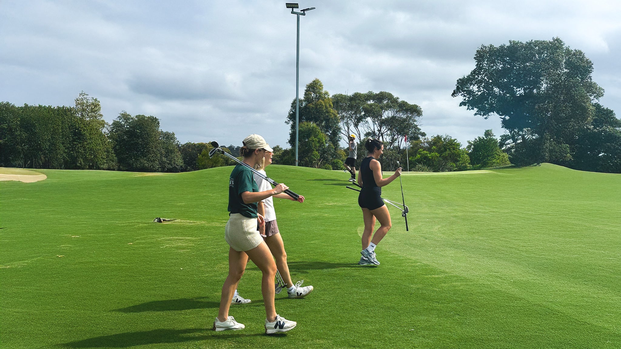 Three women playing golf on a green course with trees and a cloudy sky in the background.
