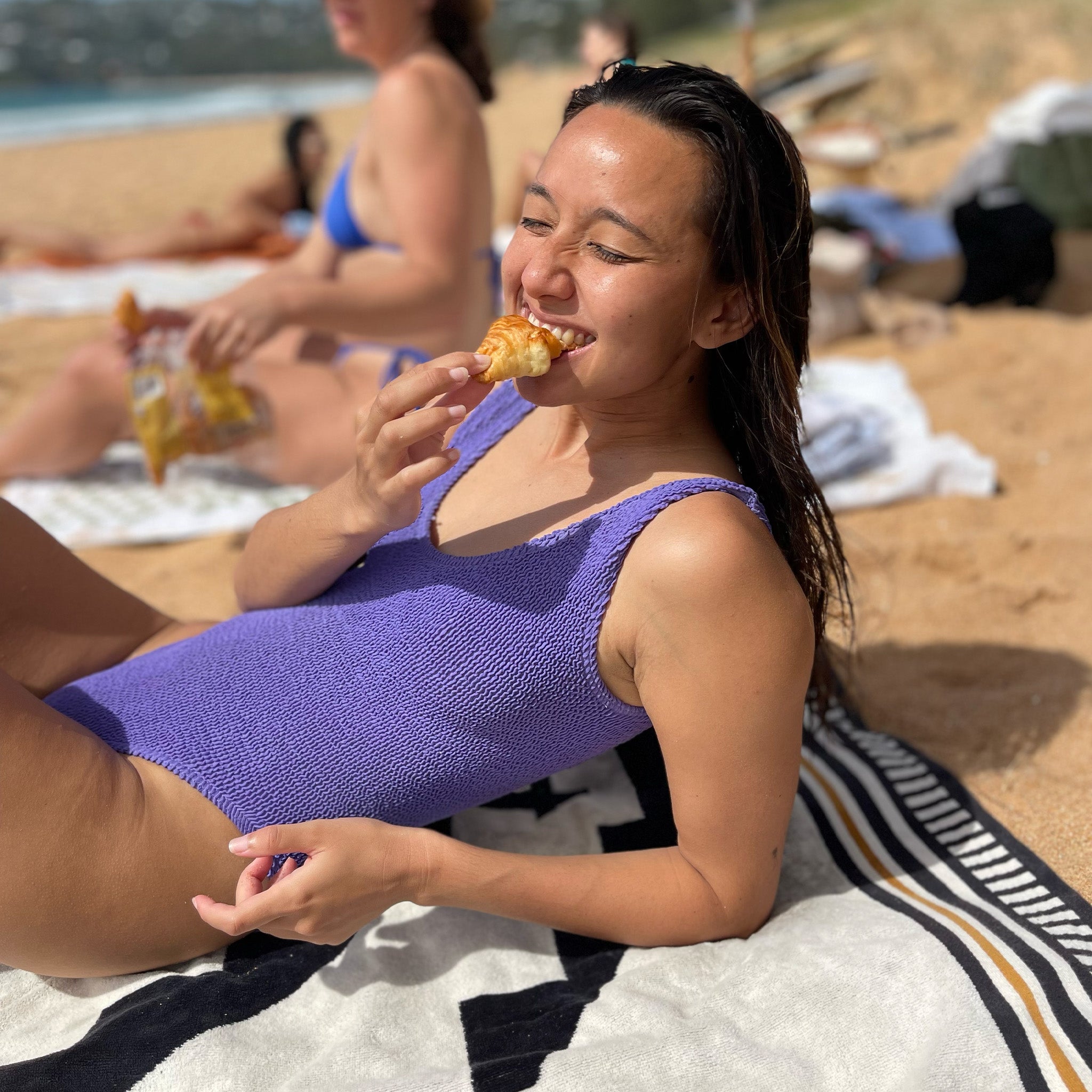 Woman in a purple swimsuit eating a croissant on a beach with other people in the background.