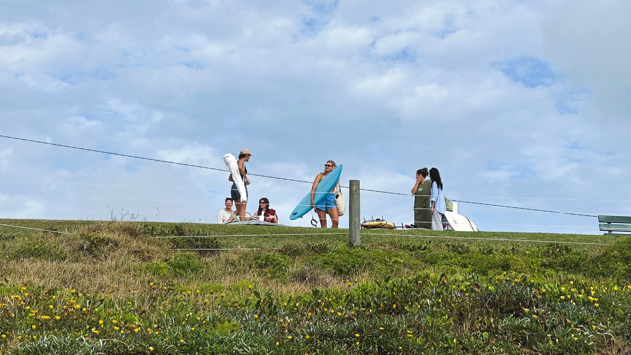 People with surfboards on a grassy hillside under a blue sky with clouds