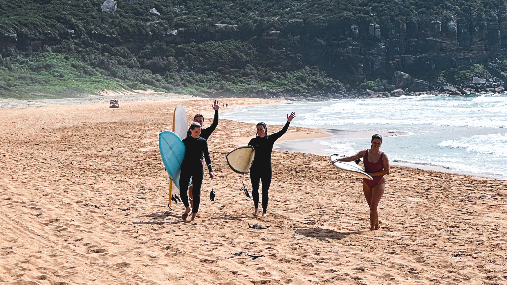 Three people with surfboards walking on a sandy beach with ocean and cliffs in the background