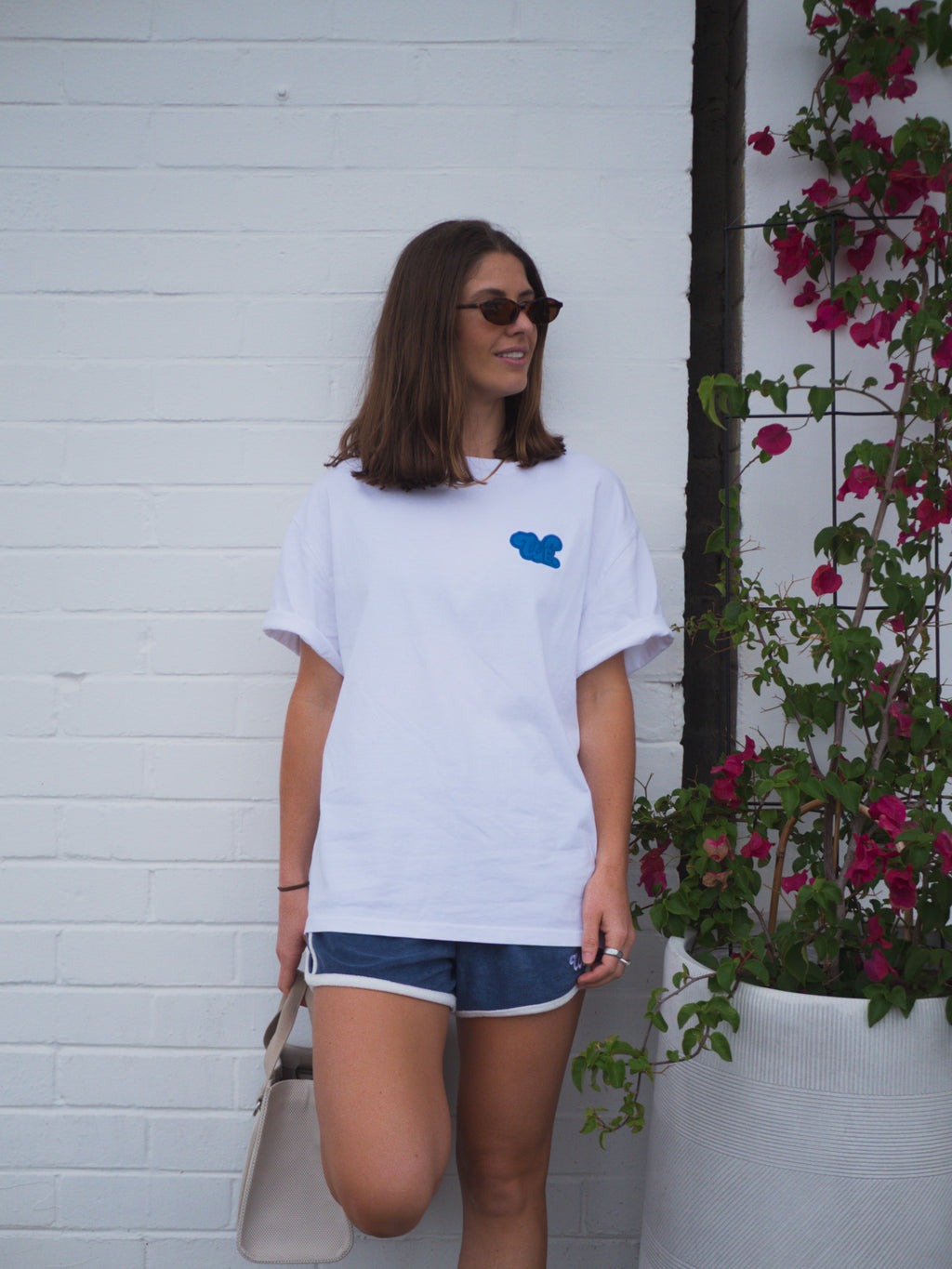 Woman wearing a white t-shirt with a blue logo, standing against a white wall with plants.