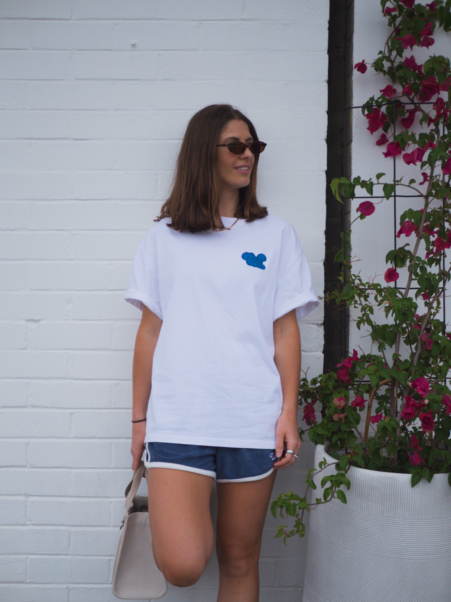 Woman wearing a white t-shirt with a blue logo, standing against a white wall with plants.