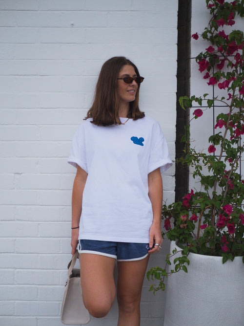 Woman wearing a white t-shirt with a blue logo, standing against a white wall with plants.